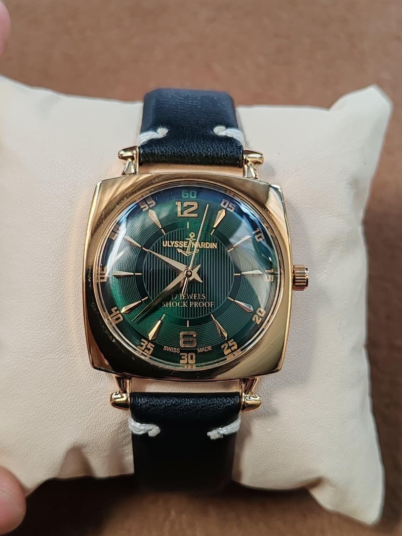 K&ouml;nnte beinhalten: Eine goldfarbene Armbanduhr mit quadratischem Geh&auml;use und dunkelgr&uuml;nem Zifferblatt mit Sonnenschliff, goldenen Indizes und Zeigern. Das Zifferblatt zeigt den Text ULYSSE NARDIN, 17 JEWELS, SHOCK PROOF und SWISS MADE. Sie hat ein schwarzes Lederarmband mit wei&szlig;en N&auml;hten.