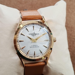 May include: A gold-tone wristwatch with a white textured dial, gold hands, and gold hour markers. The dial features the text ULYSSE NARDIN, 17 JEWELS, SHOCK PROOF, and SWISS MADE. The watch has a tan leather strap and a polished gold-tone metal case.