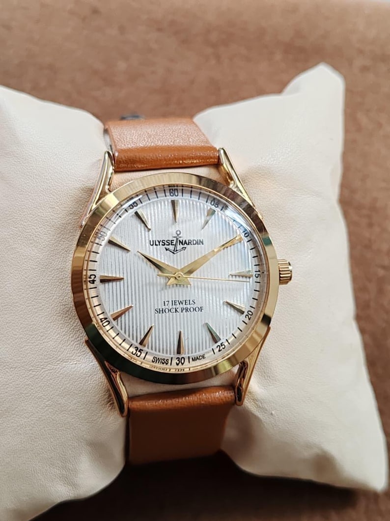 May include: A gold-tone wristwatch with a white textured dial, gold hands, and gold hour markers. The dial features the text ULYSSE NARDIN, 17 JEWELS, SHOCK PROOF, and SWISS MADE. The watch has a tan leather strap and a polished gold-tone metal case.