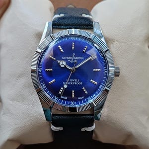 May include: A silver-tone wristwatch with a deep blue dial, featuring the text ULYSSE NARDIN, 17 JEWELS, SHOCK PROOF, and SWISS MADE. The watch has a fluted bezel, silver hands, and a black leather strap with white stitching, presented on a cushion.
