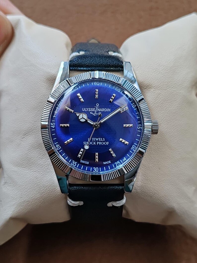 May include: A silver-tone wristwatch with a deep blue dial, featuring the text ULYSSE NARDIN, 17 JEWELS, SHOCK PROOF, and SWISS MADE. The watch has a fluted bezel, silver hands, and a black leather strap with white stitching, presented on a cushion.