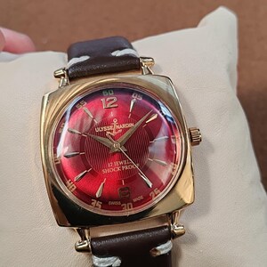May include: A gold-tone square wristwatch with a vibrant red sunburst dial featuring the Ulysse Nardin logo, an anchor symbol, and the text 17 JEWEL SHOCK PROOF. The watch has gold-tone hour markers, a brown leather strap with white stitching.