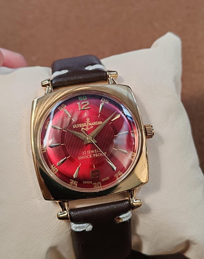 May include: A gold-tone square wristwatch with a vibrant red sunburst dial featuring the Ulysse Nardin logo, an anchor symbol, and the text 17 JEWEL SHOCK PROOF. The watch has gold-tone hour markers, a brown leather strap with white stitching.