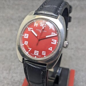 May include: A wristwatch with a silver-toned cushion-shaped case and a vibrant red dial. The dial features white Arabic numerals, white hands, and the text ULYSSE NARDIN and SWISS MADE. It is attached to a black leather strap with a textured finish.