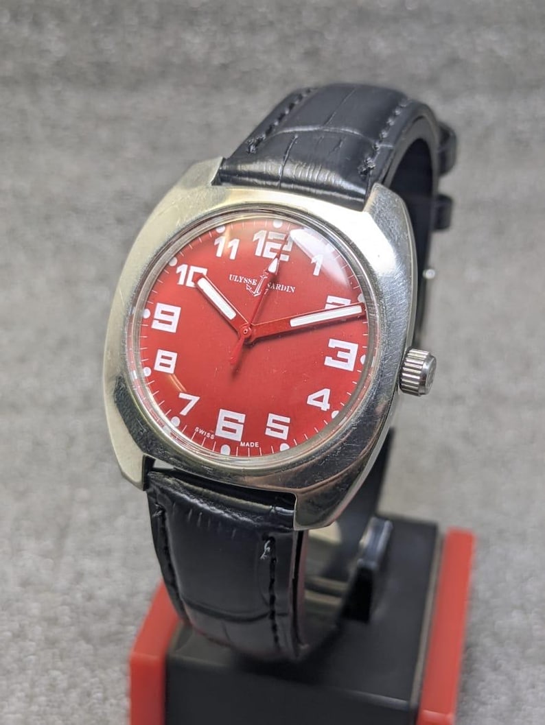 May include: A wristwatch with a silver-toned cushion-shaped case and a vibrant red dial. The dial features white Arabic numerals, white hands, and the text ULYSSE NARDIN and SWISS MADE. It is attached to a black leather strap with a textured finish.