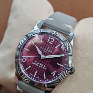 May include: A vintage Ulysse Nardin wristwatch with a deep purple dial featuring vertical stripes, silver-tone hands, and hour markers. The watch has a fluted silver-tone bezel, a stainless steel case, and a light gray leather strap with white stitching.