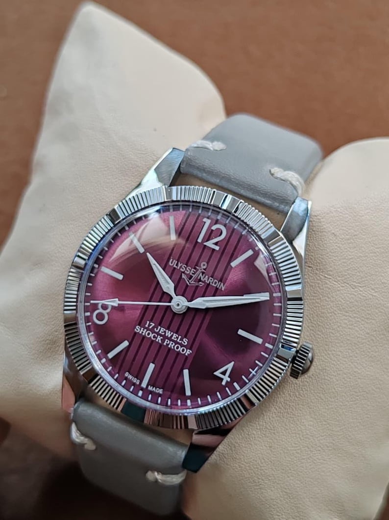 May include: A vintage Ulysse Nardin wristwatch with a deep purple dial featuring vertical stripes, silver-tone hands, and hour markers. The watch has a fluted silver-tone bezel, a stainless steel case, and a light gray leather strap with white stitching.