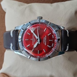 May include: A Ulysse Nardin wristwatch featuring a vibrant red sunburst dial with silver-tone hour markers, silver hands, and a fluted silver bezel. The watch includes a dark brown leather strap with white stitching and displays 17 JEWELS SHOCK PROOF.