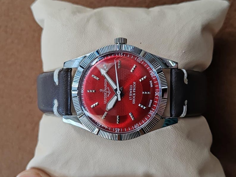 May include: A Ulysse Nardin wristwatch featuring a vibrant red sunburst dial with silver-tone hour markers, silver hands, and a fluted silver bezel. The watch includes a dark brown leather strap with white stitching and displays 17 JEWELS SHOCK PROOF.