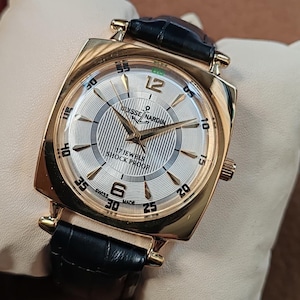 May include: A gold-tone wristwatch with a square case and a black leather strap. The silver dial features a textured center, gold-tone hands, and indices. Text on the dial reads Ulysse Nardin, 17 Jewels, Shock Proof, and Swiss Made, with numerals 6 and 12.