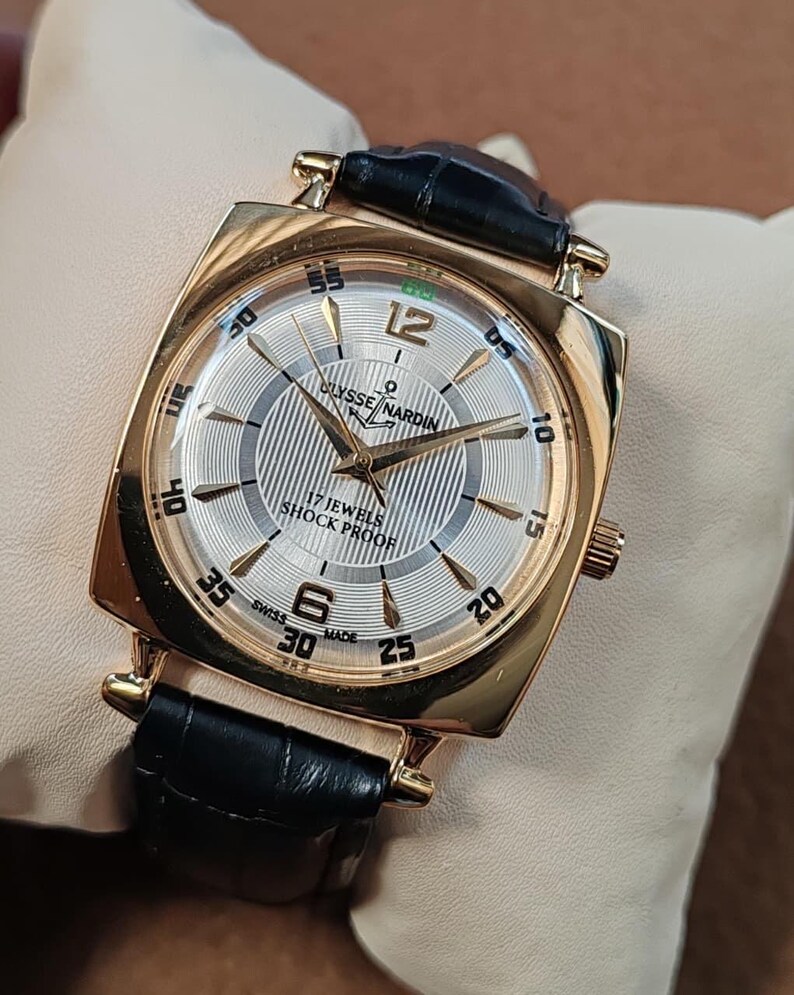 May include: A gold-tone wristwatch with a square case and a black leather strap. The silver dial features a textured center, gold-tone hands, and indices. Text on the dial reads Ulysse Nardin, 17 Jewels, Shock Proof, and Swiss Made, with numerals 6 and 12.