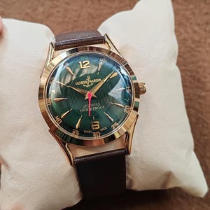 May include: A gold-tone wristwatch with a dark green dial featuring a textured pattern, gold-tone hour markers, and numerals at 12 and 6. The dial displays ULYSSE NARDIN, 17 JEWELS, SHOCK PROOF, and SWISS MADE text with a red second hand and brown strap.