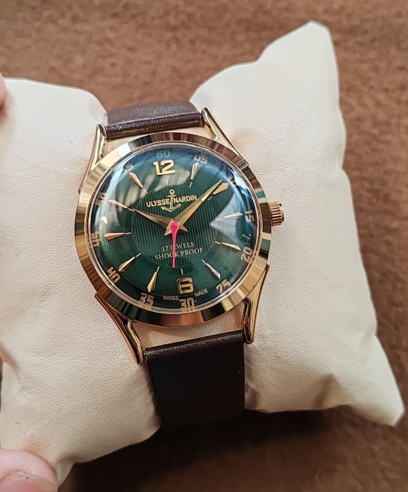 May include: A gold-tone wristwatch with a dark green dial featuring a textured pattern, gold-tone hour markers, and numerals at 12 and 6. The dial displays ULYSSE NARDIN, 17 JEWELS, SHOCK PROOF, and SWISS MADE text with a red second hand and brown strap.