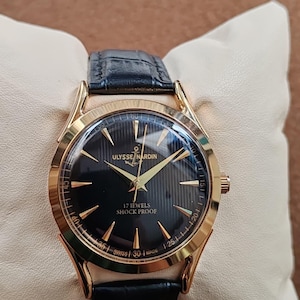 Puede incluir: Un reloj de pulsera dorado con esfera negra que presenta el texto Ulysse Nardin, 17 Jewels, Shock Proof y Swiss Made. El reloj tiene manecillas e &iacute;ndices dorados, una correa de cuero negra y una caja redonda con acabado pulido.