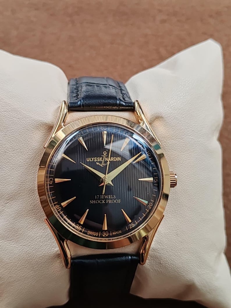 Puede incluir: Un reloj de pulsera dorado con esfera negra que presenta el texto Ulysse Nardin, 17 Jewels, Shock Proof y Swiss Made. El reloj tiene manecillas e &iacute;ndices dorados, una correa de cuero negra y una caja redonda con acabado pulido.