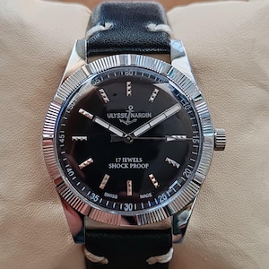 K&ouml;nnte beinhalten: Eine Armbanduhr mit schwarzem Zifferblatt, silberfarbenen Zeigern und silbernen Stundenmarkierungen. Das Zifferblatt tr&auml;gt die Aufschrift ULYSSE NARDIN, 17 JEWELS, SHOCK PROOF und SWISS MADE. Sie hat eine geriffelte silberne L&uuml;nette und ein schwarzes Lederarmband.