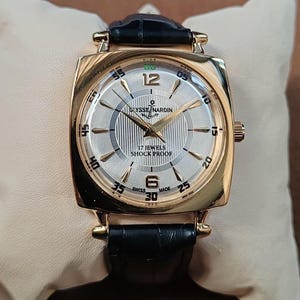 May include: A gold-tone wristwatch with a square case and a black leather strap. The silver dial features a textured center, gold-tone hands, and indices. Text on the dial reads Ulysse Nardin, 17 Jewels, Shock Proof, and Swiss Made with numerals 12 and 6.