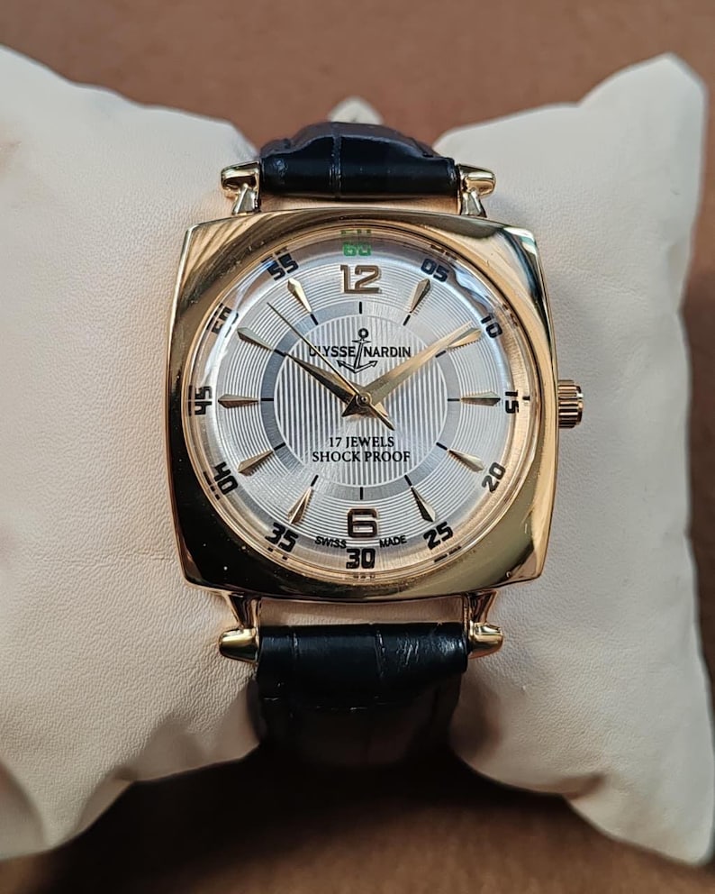 May include: A gold-tone wristwatch with a square case and a black leather strap. The silver dial features a textured center, gold-tone hands, and indices. Text on the dial reads Ulysse Nardin, 17 Jewels, Shock Proof, and Swiss Made with numerals 12 and 6.