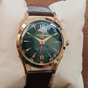 May include: A gold-tone wristwatch with a dark green dial, featuring the text Ulysse Nardin, 17 Jewels, Shock Proof, and Swiss Made. The watch has gold-tone hour markers, a red second hand, and a brown leather strap with a polished gold-tone case.