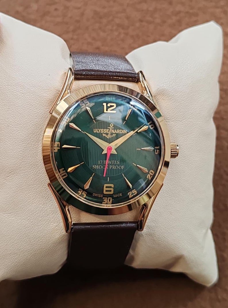 May include: A gold-tone wristwatch with a dark green dial, featuring the text Ulysse Nardin, 17 Jewels, Shock Proof, and Swiss Made. The watch has gold-tone hour markers, a red second hand, and a brown leather strap with a polished gold-tone case.