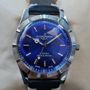May include: A wristwatch with a circular blue dial, silver-tone hands, and silver-tone hour markers. The dial features the text ULYSSE NARDIN, an anchor logo, 17 JEWELS, SHOCK PROOF, and SWISS MADE. It has a fluted silver bezel and a black leather strap.