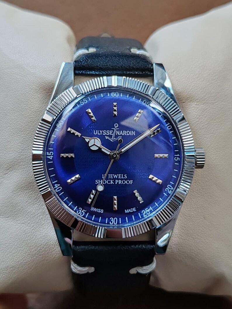 May include: A wristwatch with a circular blue dial, silver-tone hands, and silver-tone hour markers. The dial features the text ULYSSE NARDIN, an anchor logo, 17 JEWELS, SHOCK PROOF, and SWISS MADE. It has a fluted silver bezel and a black leather strap.