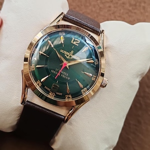 May include: A gold-tone wristwatch with a dark green dial, featuring gold-tone hour markers and hands, a red second hand, and a brown leather strap. The dial displays the text Ulysse Nardin, 17 Jewels, Shock Proof, and Swiss Made.