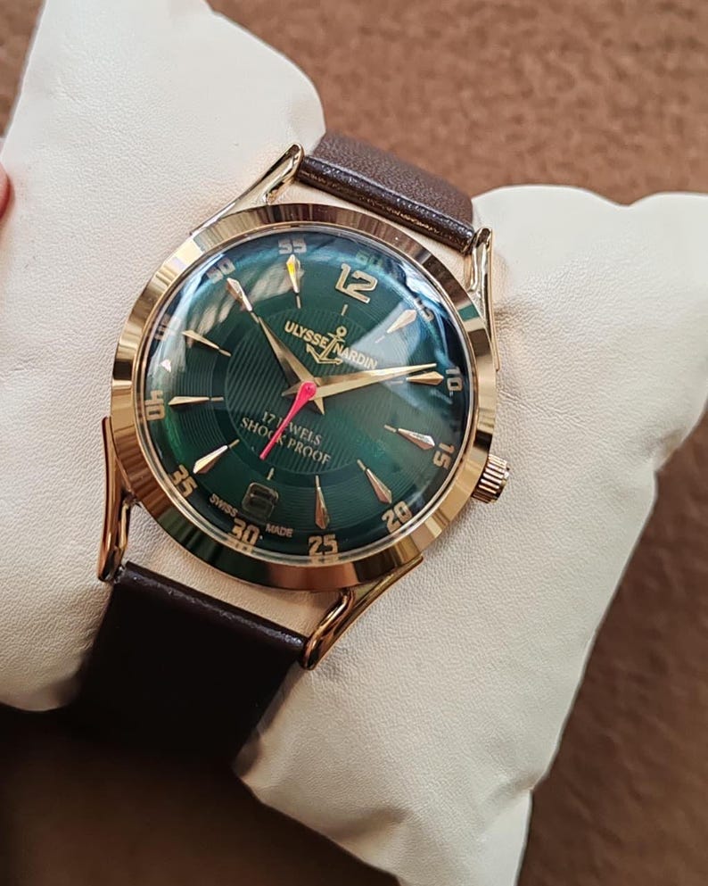 May include: A gold-tone wristwatch with a dark green dial, featuring gold-tone hour markers and hands, a red second hand, and a brown leather strap. The dial displays the text Ulysse Nardin, 17 Jewels, Shock Proof, and Swiss Made.