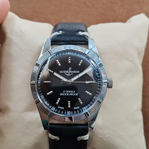 K&ouml;nnte beinhalten: Eine Armbanduhr mit schwarzem Zifferblatt, silberfarbenen Zeigern und Indizes. Das Zifferblatt tr&auml;gt die Aufschrift Ulysse Nardin, 17 Jewels, Shock Proof und Swiss Made. Sie hat eine geriffelte silberfarbene L&uuml;nette und ein schwarzes Lederarmband.