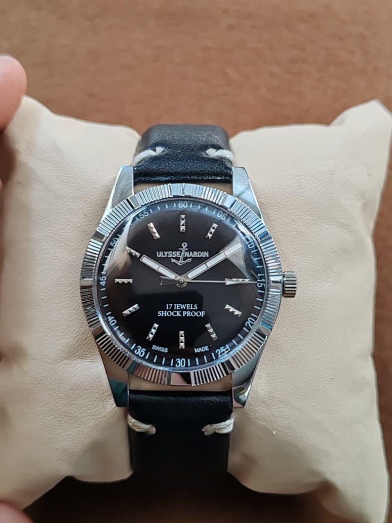 K&ouml;nnte beinhalten: Eine Armbanduhr mit schwarzem Zifferblatt, silberfarbenen Zeigern und Indizes. Das Zifferblatt tr&auml;gt die Aufschrift Ulysse Nardin, 17 Jewels, Shock Proof und Swiss Made. Sie hat eine geriffelte silberfarbene L&uuml;nette und ein schwarzes Lederarmband.
