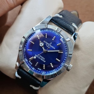 May include: A Ulysse Nardin wristwatch with a deep blue sunburst dial, silver-tone hands, and hour markers. The watch features a fluted silver bezel, a black leather strap with white stitching, and text reading Ulysse Nardin, Jewels, and Shock Proof.