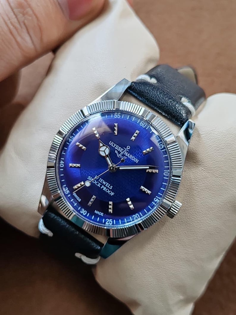 May include: A Ulysse Nardin wristwatch with a deep blue sunburst dial, silver-tone hands, and hour markers. The watch features a fluted silver bezel, a black leather strap with white stitching, and text reading Ulysse Nardin, Jewels, and Shock Proof.