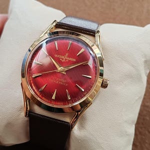 May include: A gold-tone wristwatch with a vibrant red dial featuring a textured striped pattern. The dial displays the Ulysse Nardin logo, an anchor symbol, 17 Jewels, Shock Proof, and Swiss Made text. It has gold hands and a dark brown leather strap.