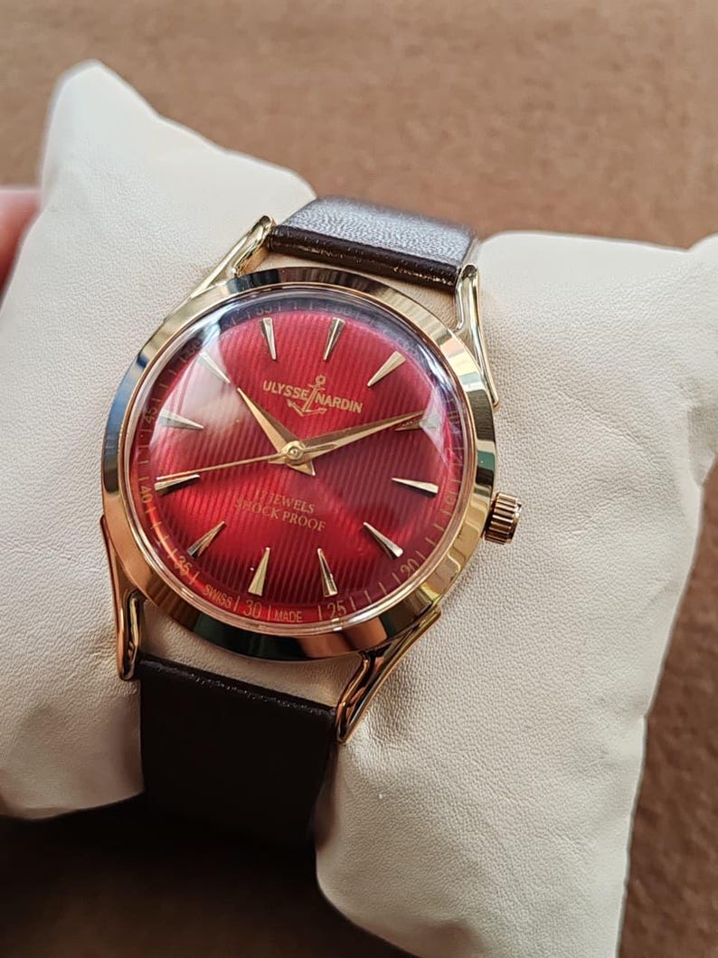 May include: A gold-tone wristwatch with a vibrant red dial featuring a textured striped pattern. The dial displays the Ulysse Nardin logo, an anchor symbol, 17 Jewels, Shock Proof, and Swiss Made text. It has gold hands and a dark brown leather strap.