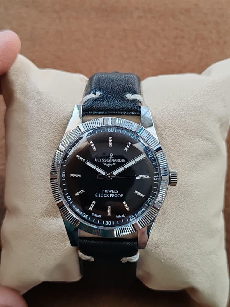 K&ouml;nnte beinhalten: Eine Ulysse Nardin Armbanduhr mit schwarzem Zifferblatt, silberfarbenen Zeigern und Stundenmarkierungen. Die Uhr verf&uuml;gt &uuml;ber eine geriffelte silberne L&uuml;nette, ein schwarzes Lederarmband mit wei&szlig;en N&auml;hten und den Schriftzug Ulysse Nardin, 17 Jewels, Shock Proof, Swiss Made.