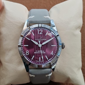 May include: A wristwatch with a silver-toned fluted bezel and a deep purple dial featuring vertical stripes. The dial displays the text ULYSSE NARDIN, 17 JEWELS, SHOCK PROOF, and SWISS MADE, with white numerals at 12, 8, and 4, and a grey leather strap.