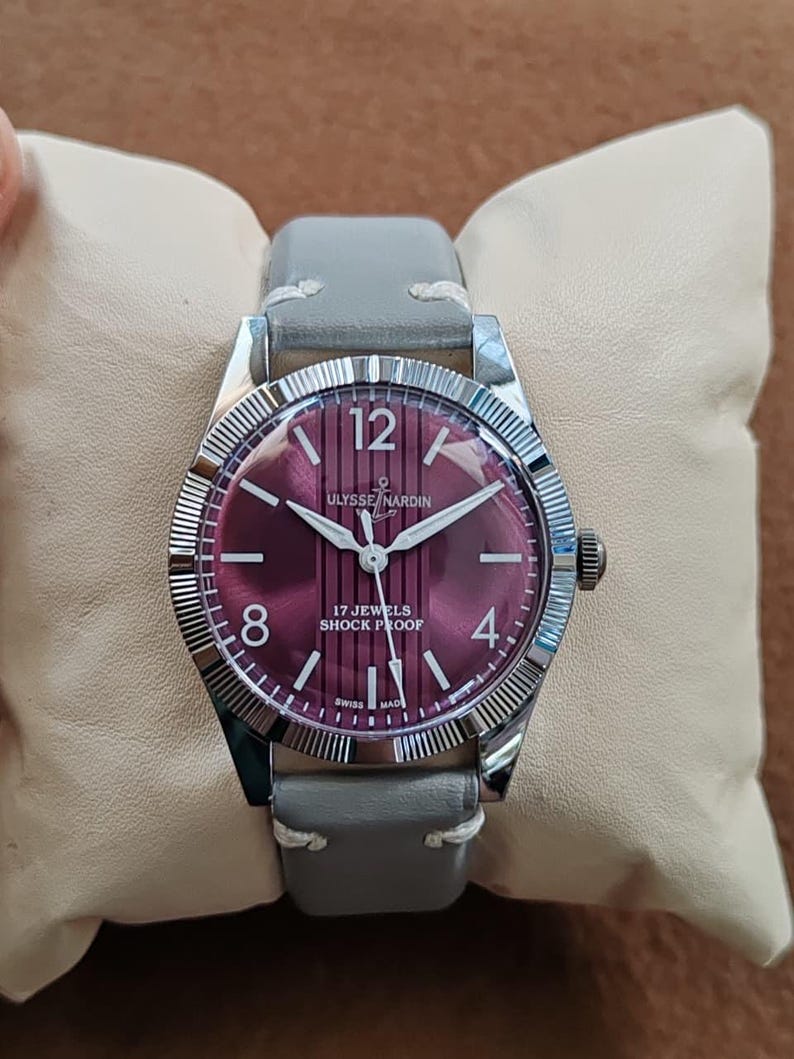 May include: A wristwatch with a silver-toned fluted bezel and a deep purple dial featuring vertical stripes. The dial displays the text ULYSSE NARDIN, 17 JEWELS, SHOCK PROOF, and SWISS MADE, with white numerals at 12, 8, and 4, and a grey leather strap.