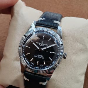 K&ouml;nnte beinhalten: Eine silberfarbene Armbanduhr mit schwarzem Zifferblatt, das die Aufschrift ULYSSE NARDIN, 17 JEWELS, SHOCK PROOF und SWISS MADE tr&auml;gt. Die Uhr verf&uuml;gt &uuml;ber eine geriffelte L&uuml;nette, silberne Zeiger und ein schwarzes Lederarmband mit wei&szlig;en N&auml;hten.