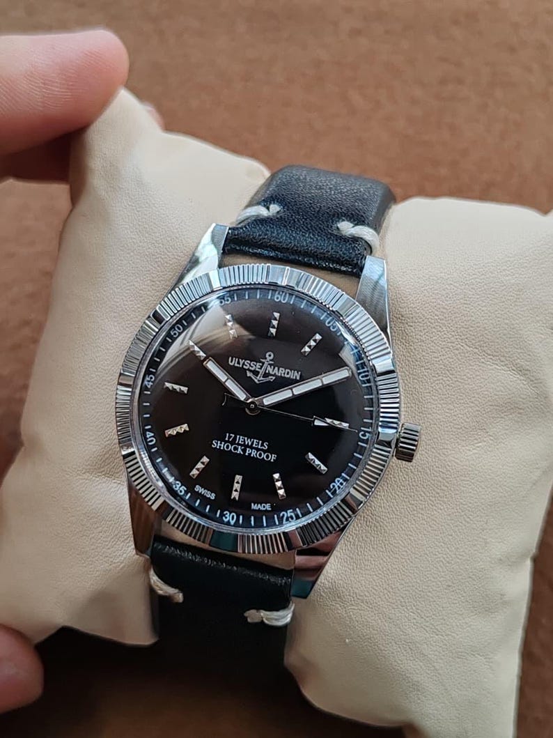 K&ouml;nnte beinhalten: Eine silberfarbene Armbanduhr mit schwarzem Zifferblatt, das die Aufschrift ULYSSE NARDIN, 17 JEWELS, SHOCK PROOF und SWISS MADE tr&auml;gt. Die Uhr verf&uuml;gt &uuml;ber eine geriffelte L&uuml;nette, silberne Zeiger und ein schwarzes Lederarmband mit wei&szlig;en N&auml;hten.