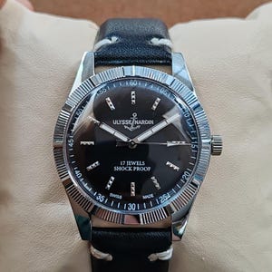 K&ouml;nnte beinhalten: Eine Ulysse Nardin Armbanduhr mit schwarzem Zifferblatt, silberfarbenen Zeigern und Stundenmarkierungen. Die Uhr verf&uuml;gt &uuml;ber eine geriffelte silberne L&uuml;nette, ein schwarzes Lederarmband mit wei&szlig;en N&auml;hten und die Aufschrift Ulysse Nardin, 17 Jewels, Shock Proof, Swiss Made.