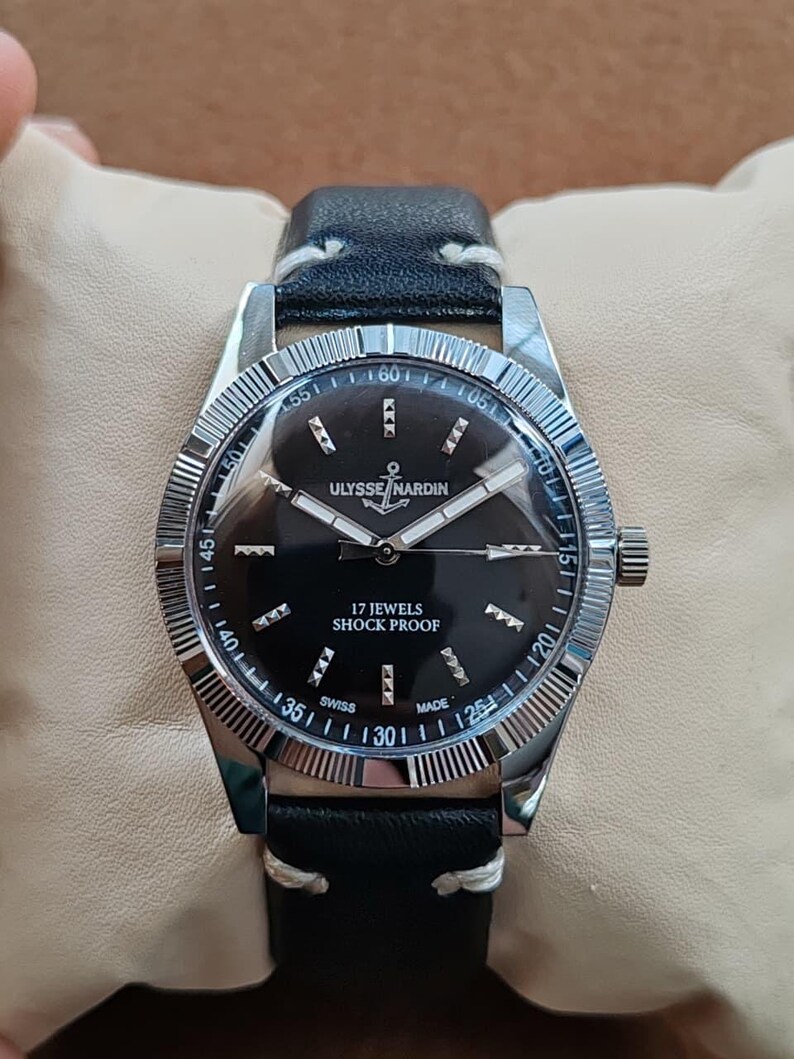 K&ouml;nnte beinhalten: Eine Ulysse Nardin Armbanduhr mit schwarzem Zifferblatt, silberfarbenen Zeigern und Stundenmarkierungen. Die Uhr verf&uuml;gt &uuml;ber eine geriffelte silberne L&uuml;nette, ein schwarzes Lederarmband mit wei&szlig;en N&auml;hten und die Aufschrift Ulysse Nardin, 17 Jewels, Shock Proof, Swiss Made.