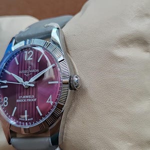 May include: A wristwatch with a round, deep purple dial featuring vertical stripes, silver-tone hands, and indices. The dial displays the text ULYSSE NARDIN, 17 JEWELS, SHOCK PROOF, and SWISS MADE. It has a silver-tone fluted bezel and a grey leather strap.