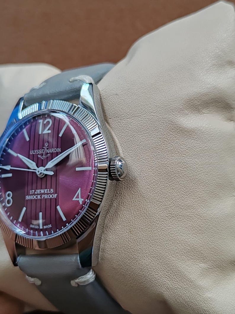 May include: A wristwatch with a round, deep purple dial featuring vertical stripes, silver-tone hands, and indices. The dial displays the text ULYSSE NARDIN, 17 JEWELS, SHOCK PROOF, and SWISS MADE. It has a silver-tone fluted bezel and a grey leather strap.