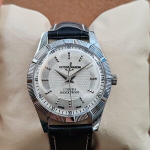 May include: A silver Ulysse Nardin wristwatch with a black leather band. The watch face is silver with black hands and hour markers. The watch has the text "17 JEWELS SHOCK PROOF" and "SWISS MADE". The watch is on a cream-colored pillow.