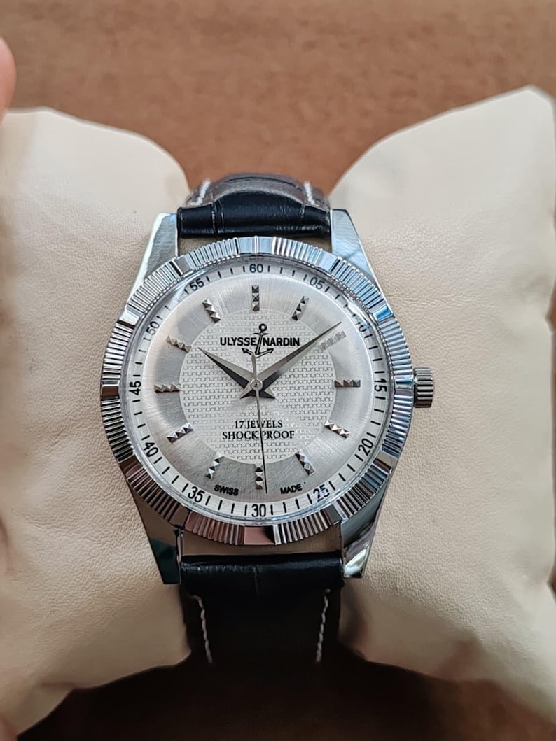 May include: A silver Ulysse Nardin wristwatch with a black leather band. The watch face is silver with black hands and hour markers. The watch has the text "17 JEWELS SHOCK PROOF" and "SWISS MADE". The watch is on a cream-colored pillow.