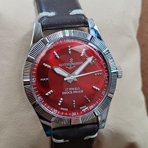 May include: A wristwatch with a dark brown leather strap and a silver-toned fluted bezel. The watch face is a deep red with silver hour markers, silver hands, and the text ULYSSE NARDIN, 17 JEWELS, SHOCK PROOF, and SWISS MADE printed in white.