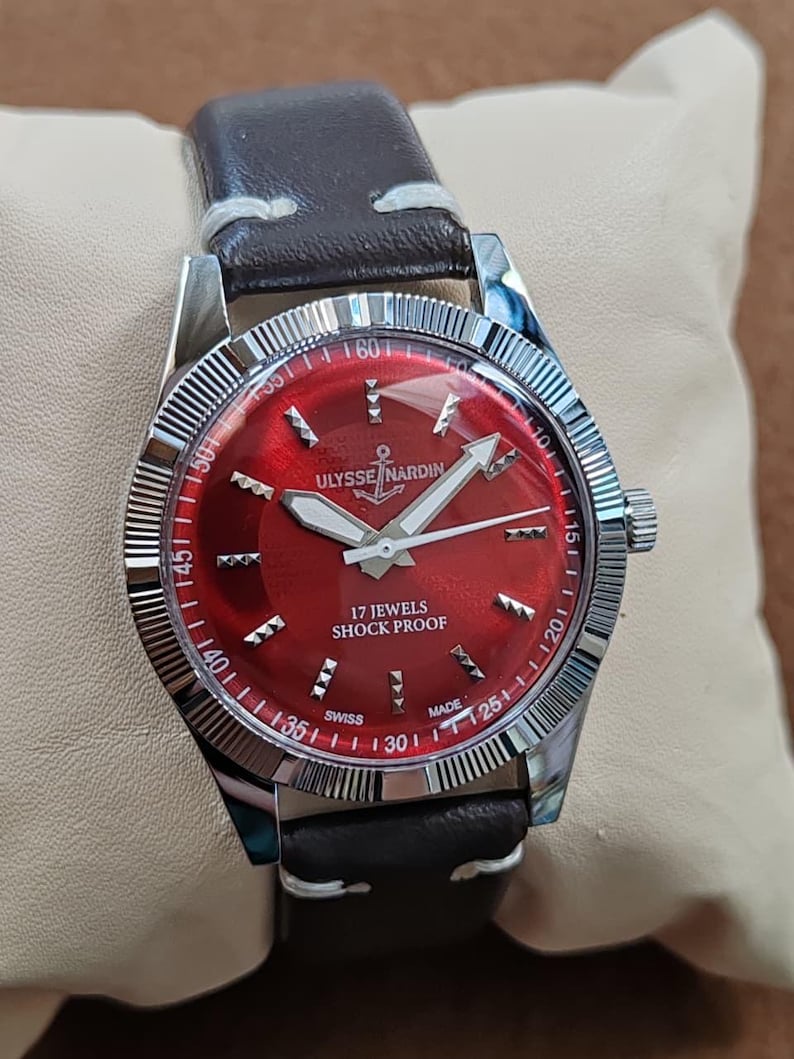 May include: A wristwatch with a dark brown leather strap and a silver-toned fluted bezel. The watch face is a deep red with silver hour markers, silver hands, and the text ULYSSE NARDIN, 17 JEWELS, SHOCK PROOF, and SWISS MADE printed in white.
