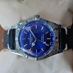 May include: A Ulysse Nardin wristwatch featuring a round silver-tone case with a fluted bezel and a vibrant blue sunburst dial. The dial displays silver-tone hour markers, an anchor logo, and the text Ulysse Nardin, 17 Jewels, and Shock Proof.
