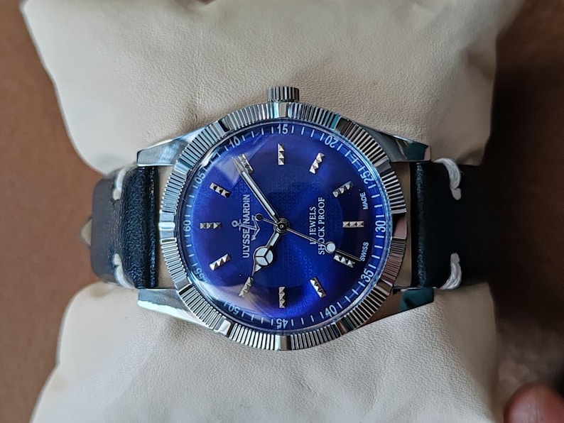 May include: A Ulysse Nardin wristwatch featuring a round silver-tone case with a fluted bezel and a vibrant blue sunburst dial. The dial displays silver-tone hour markers, an anchor logo, and the text Ulysse Nardin, 17 Jewels, and Shock Proof.