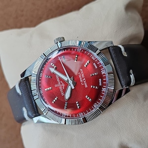 May include: A wristwatch with a vibrant red dial, silver-tone hands, and hour markers. The face features the text Ulysse Nardin, 17 Jewels, and Shock Proof. It has a silver fluted bezel and a dark brown leather strap with white stitching.